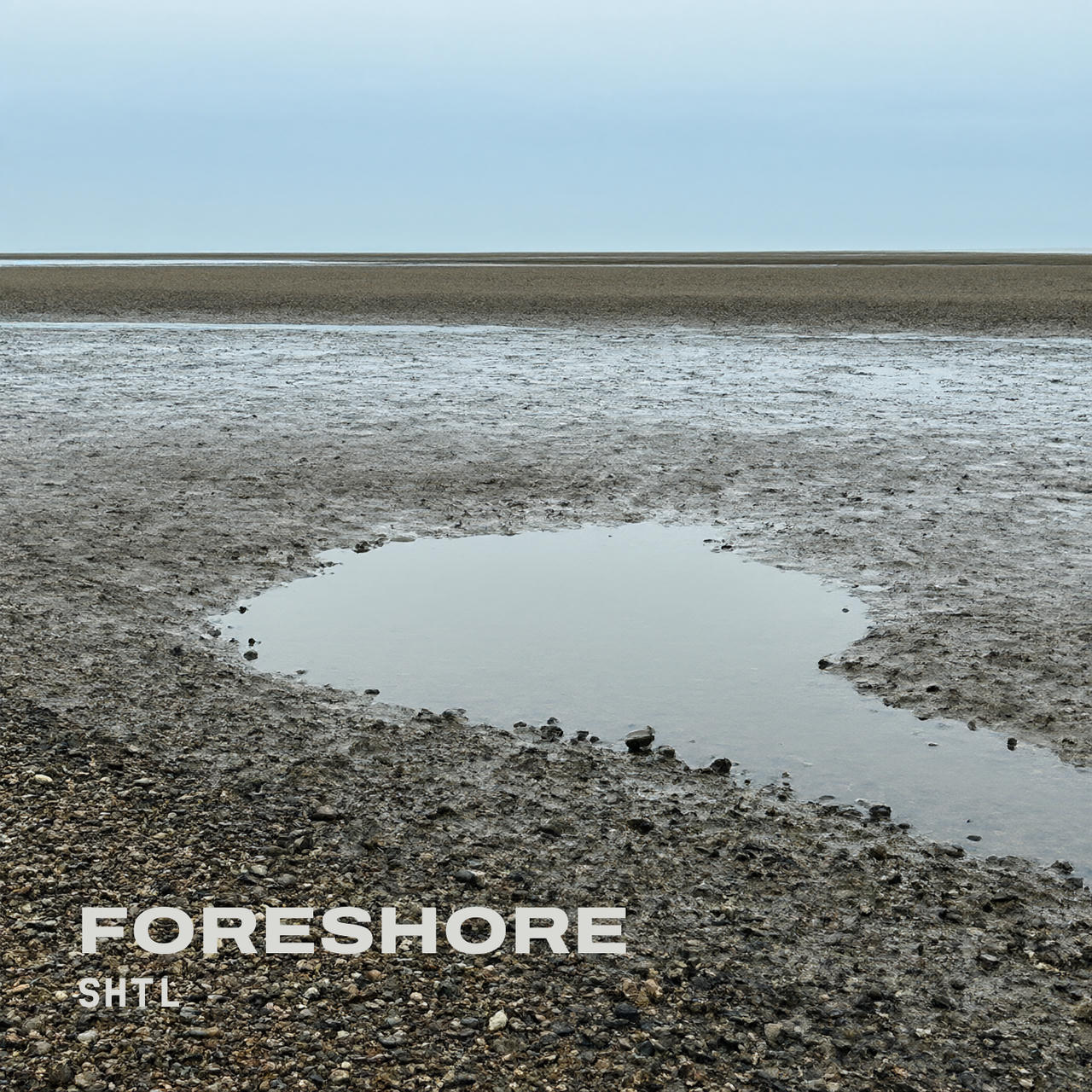 A wide, flat foreshore at low tide under a pale grey-blue sky, with wet sand and shingle stretching to a thin line of sea on the horizon and a shallow pool of still water in the middle ground. In the lower left corner, "FORESHORE" is written in large bold off-white capitals, with "SHTL" in smaller spaced letters beneath.