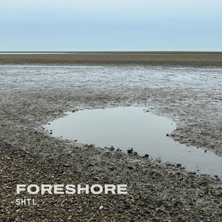 A wide, flat foreshore at low tide under a pale grey-blue sky, with wet sand and shingle stretching to a thin line of sea on the horizon and a shallow pool of still water in the middle ground. In the lower left corner, "FORESHORE" is written in large bold off-white capitals, with "SHTL" in smaller spaced letters beneath.